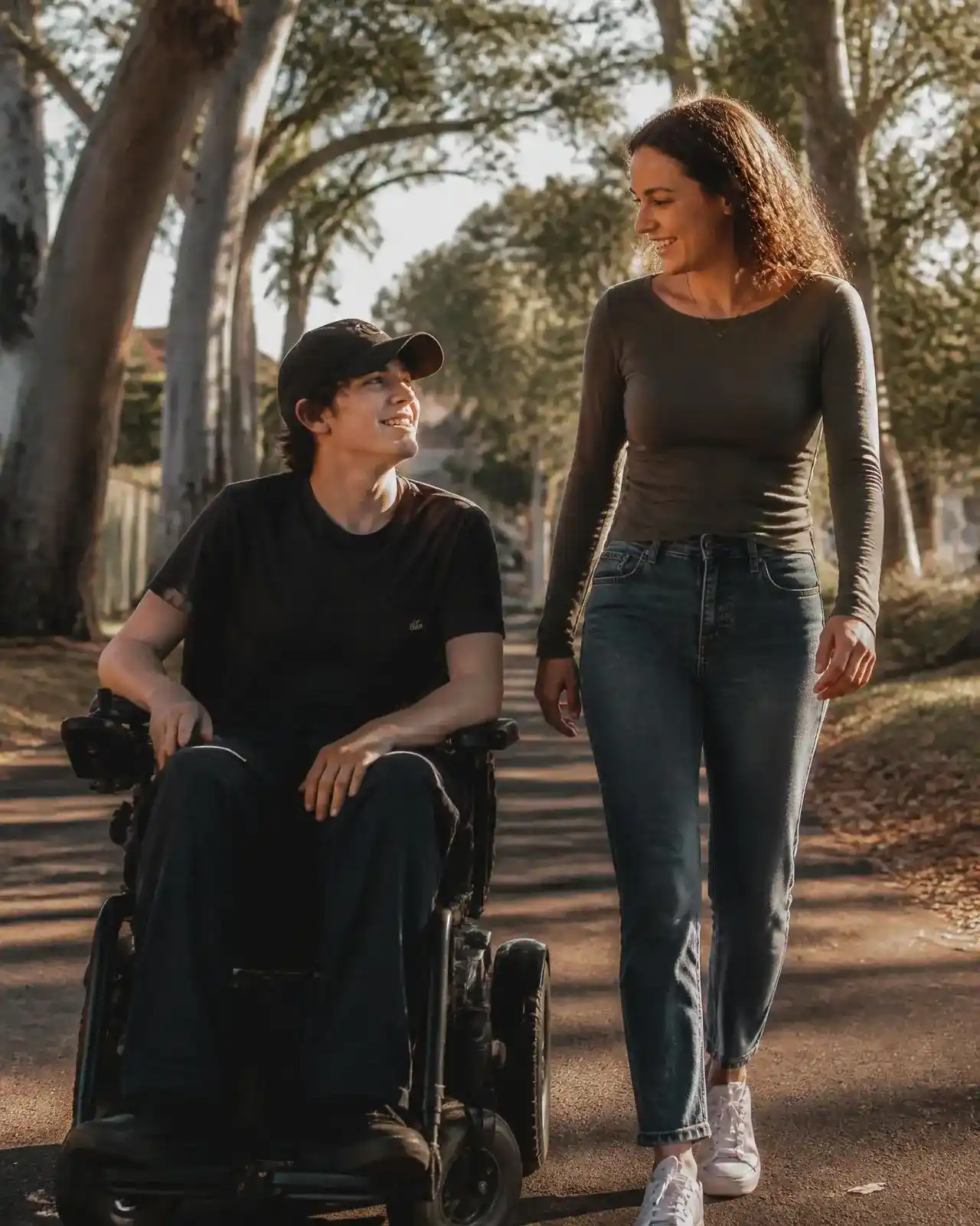 Emma, a Support Network worker, walking and chatting with a young man who uses a power wheelchair, on a sunlit Australian street lined with eucalyptus trees